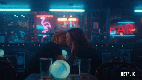 A GIF from Mae Martin's show Feel Good. Mae's character Mae kisses George, played by co-star Charlotte Richie. They are sat in a dimly lit bar with blue lights. 