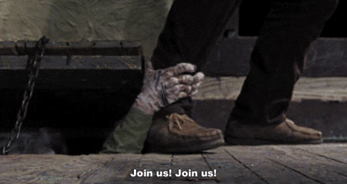 A zombie-like person reaches from beneath a wooden trapdoor and grabs a man's leg and attempts to pull him in as he tries to walk away.