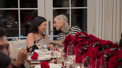 The gif of Kourtney Cardashian and Travis Barker kissing. They sit at the head of the table. there are glasses and plates and red roses on the table.