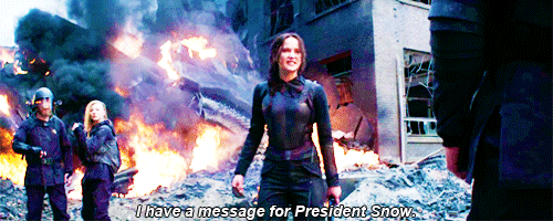 Katniss Everdeen stands with her bow in hand in her Mockingjay suit in front of a burning hospital, shouting "I have a message for President Snow." Gif from media.giphy.org 