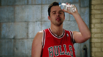 Man (character Nick Miller from TV Show New Girl) wearing a Chicago Bulls basketball singlet lifting a water bottle towards his face and making tongue movements.