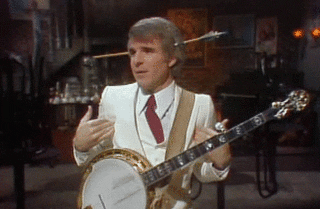 Steve Martin shrugging with a fake arrow in his head and a banjo on his lap.