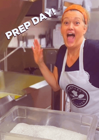 Smiling woman in an apron in a kitchen waving at the camera with the caption Prep day