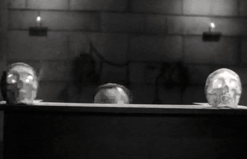 GIF of a man peaking over a counter, looking around scared. On either side of him on the counter are skulls with lights in their eyes. It is an old, black and white horror movie.
