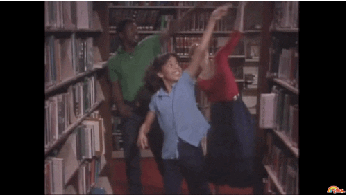 Three kids dance in sync through the stacks of a library.