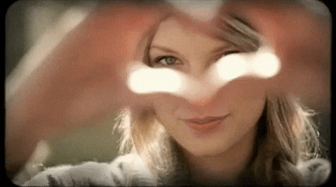 Taylor Swift holding her hands up in a heart shape and then pointing at the camera.