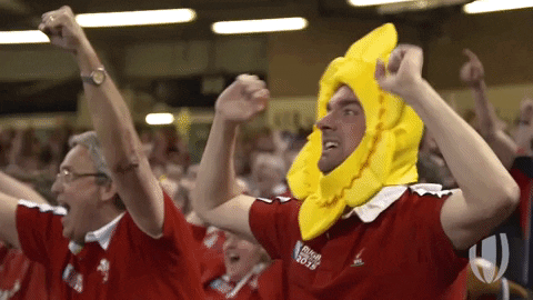 A man in a rugby uniform wears a daffodil hat. He raises his arms and yells "Yeah!"
