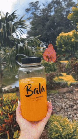 A person holds a clear bottle labeled "Bala Enzyme Bala Total Body Wellness Drink Mix" filled with orange liquid, promoting plant-based hydration, with a garden featuring trees and plants in the background.