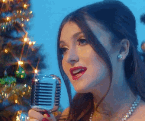 A woman sings into a microphone, standing in front of Christmas trees.