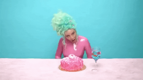 Gif of Doja Cat dancing. She wears a pink latex costume. She has turquoise hair. The background is turquoise. She has a table in front of her, a pink cake and a glass of water with goldfish on it.