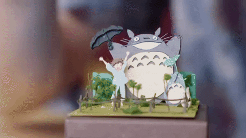 Hände platzieren vorsichtig mit einer Pinzette einen Papierausschnitt von Mei auf einem Miniaturdiorama aus dem Ghibli-Film „Mein Nachbar Totoro“.